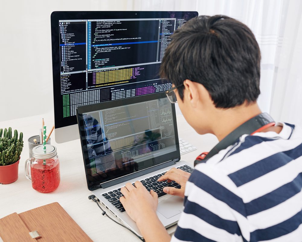 Genious teenager working on laptop, programming code on computer screen in background